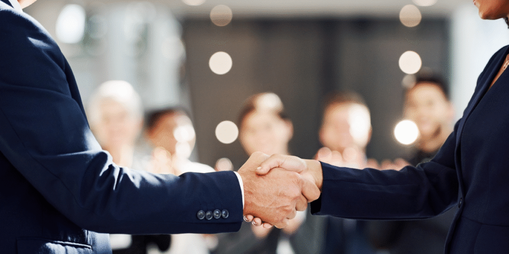 Two business professionals shaking hands, with a group of people observing in the background, symbolizing collaboration and networking.
